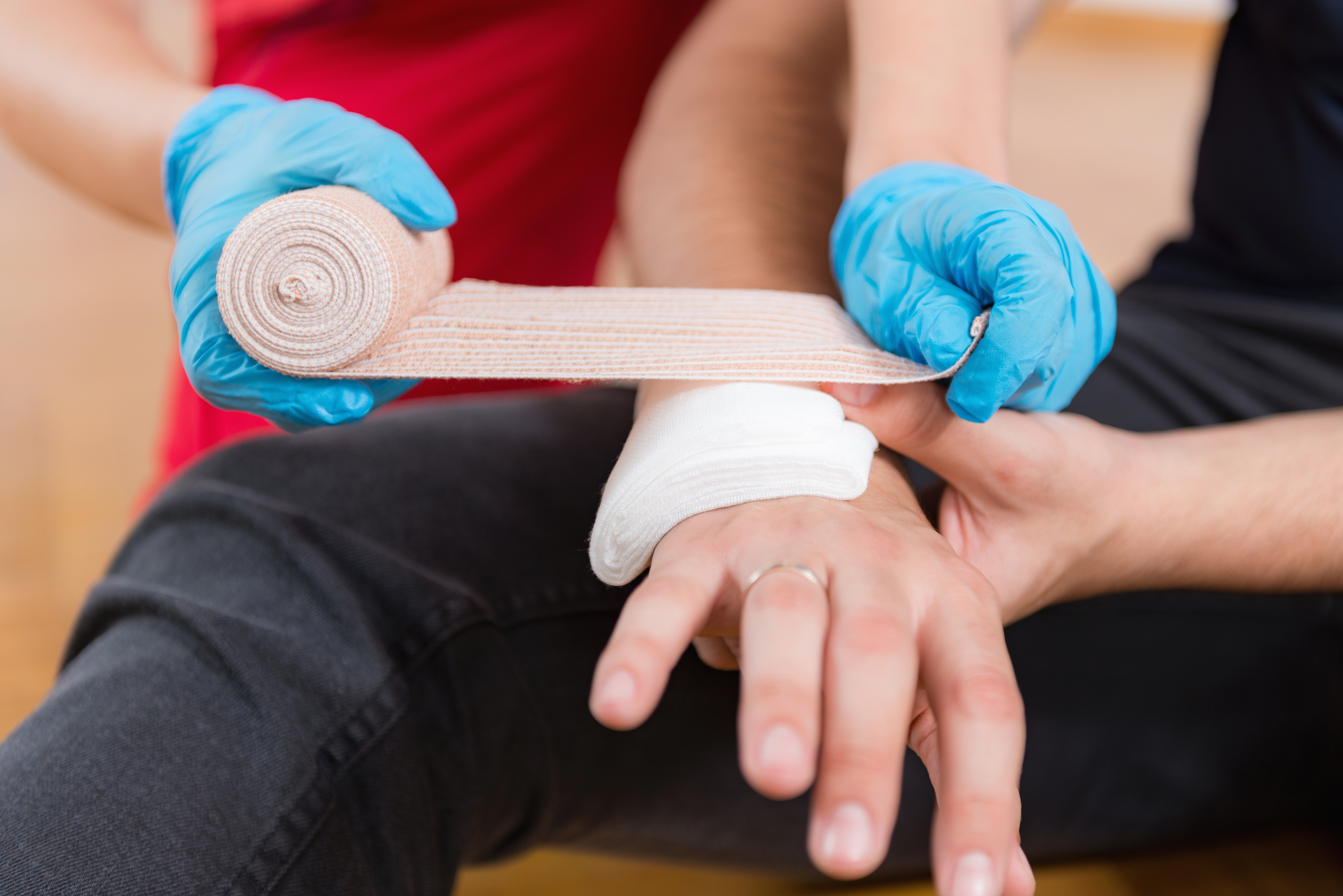 Einer Person wird von einer weiteren Person mit medizinischen Handschuhen, die rechte Hand  mit einem Verband und einer sterilen Kompresse verbunden.