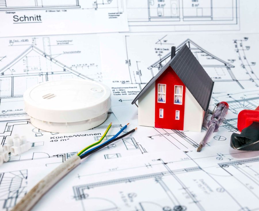 Ein Miniaturhaus steht mit einem Feuermelder und Stromkabel auf einem Hausplan.